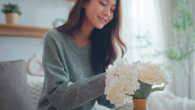 Happy Asian woman puts in vase bunch of beautiful fresh natural flowers in modern cozy living room, create cosiness at home concept