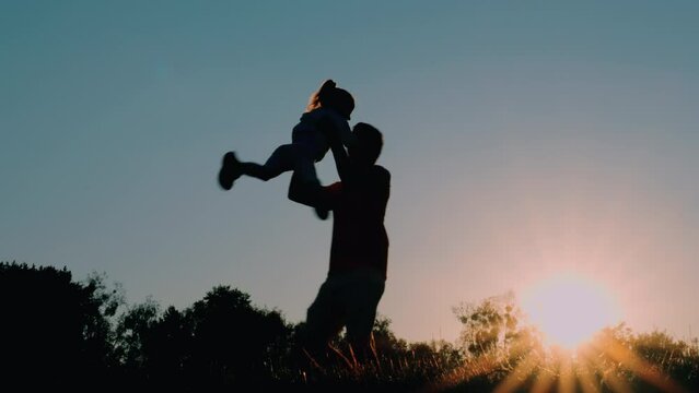 silhouettes of father and child in the evening in the meadow in the sunlight, father twirling his daughter in his arms, happy family