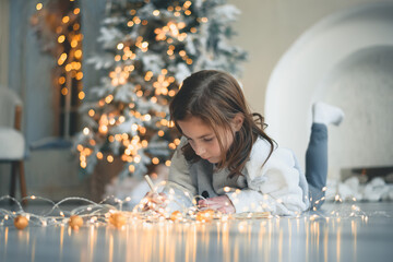 a cute girl lies on the floor near the Christmas tree and writes a letter to Santa Claus