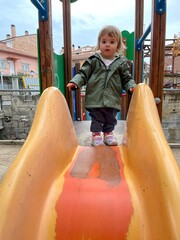 A beautiful one-year-old baby plays on a slide in a park on a cloudy day