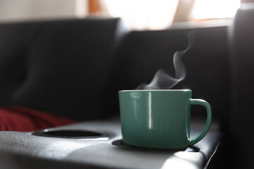 Heissgetraenk mit Dampf in türkiser Tasse auf schwarzer Couch mit Fensterlicht im Hintergrund und rotem Kissen