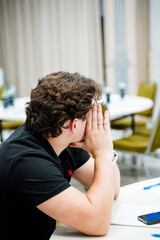 A man is sitting at a desk with his hands covering his face, a man is sitting in an office. The young man turned his head to the side.