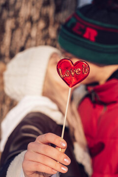 Blurred young couple in love kissing on a date on a winter day.In the foreground is a red lollipop in the shape of a heart and the inscription Love is.Valentine's day concept.