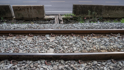 A railroad track with steel on both sides to allow trains to run. The floor is full of stones. On the side is a road. There is a small opening for people to pass through.