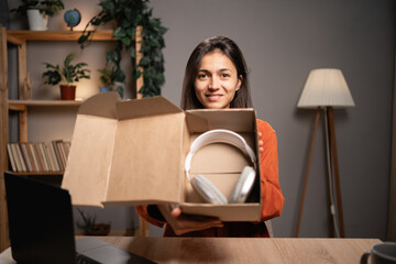 Hispanic woman opening box with ordered products. Female blogger review headphones while recording...