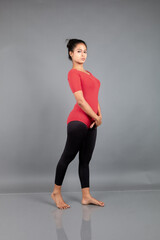 Healthy young woman in red t-shirt and black leggings doing yoga and stretching exercises isolated on grey background