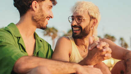 Romantic moments, homosexual couple enjoying communication with each other while sitting on the beach at sunrise, crossing their arms and looking at each other with love