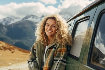 Young beautiful smiling blonde woman with curly hair standing near retro minivan in the mountains