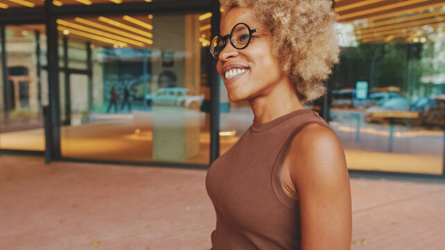 Happy Young Woman In Glasses Wearing Brown Top In Wireless Headphones Dancing Outdoors In City Street Having Fun Alone