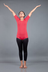 Healthy young woman in red t-shirt and black leggings doing yoga and stretching exercises isolated on grey background