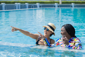 Two businesswoman friends wearing bikinis swimwear and hats with eyeglasses talking spending time together while sunbathing, healthy African and Hispanic females happy relaxing summer holiday vacation