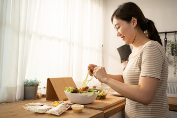 Happy smiling young Asian woman is preparing a fresh healthy vegan salad with many vegetables in...