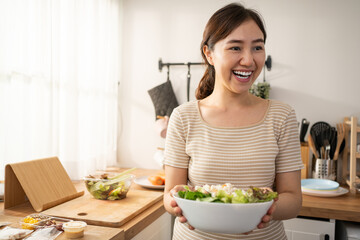 Obraz na płótnie Canvas Happy smiling young Asian woman is preparing a fresh healthy vegan salad with many vegetables in the kitchen at home and trying a new recipe.