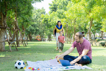 Cute little boy son healthy running with mother in park in summer holiday, Caucasian father uses modern laptop wireless technology remotely working outdoors and play with family bonding relationship