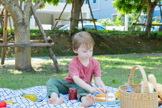 Happy Little Boy Having Fun Licking Jam In The Jar And Apply To Bread Making Sandwiches By Himself In Park Spending Time Picnic With Mom In Summer Holiday, Caucasian Family Relaxing Vacation Outdoors