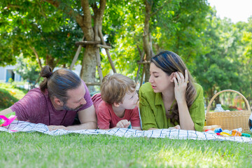 Fototapeta premium ็Hispanic or Caucasian family outdoor activities lifestyle, Mother, father and little boy lying down on grass happy kissing and embracing with love and tender, toddler playing with mom and dad in park
