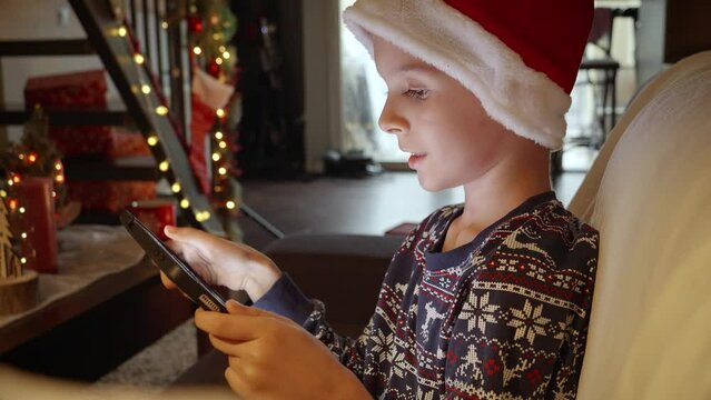 Portrait of smiling boy in Santa's hat playing games on tablet computer in house decorated for Christmas. Winter holidays, celebrations and party