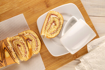 One sliced sweet cherry roll with paper plate and white cup with saucer on wooden table, top view.