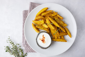 Baked potato wedges with sour cream sauce on a light background, top view. Rustic baked potatoes in the oven on a white plate with herbs.