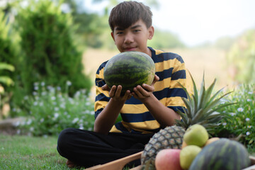 Fototapeta premium Asian boy holds watermalon sitting near fruits basket, blurred mountain background, concept for plants and fruits with children around the world.