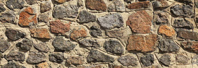 Granite wall, background. Surface of stone structure built of granite rocks (bricks). The wall texture of volcanic yellow and brown color. Fence with large stones on cement base in the rays of the sun