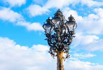 Fototapeta na wymiar Street lamp against the blue sky with clouds . Lamp base features elaborate scrolls and curves, characteristic of the Baroque period