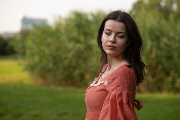 Portrait of a beautiful brunette girl in dress posing in the park, tree background. Horizontal view.
