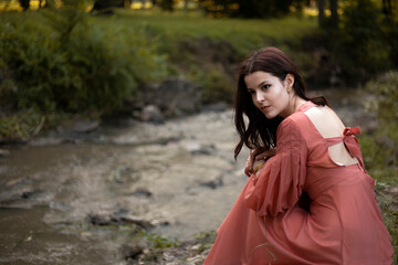 Portrait of a beautiful brunette girl in dress posing in the park, staying near river background.
