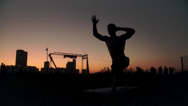 A dark male silhouette greets the sun outdoors. A man raises his hands up while doing yoga in nature. Yogi in asanas in a field, against the backdrop of the city at sunset