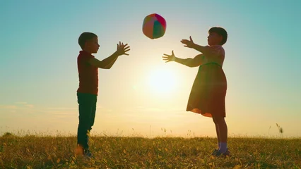 Foto op Plexiglas Children play with big ball in park against backdrop of sun. Happy child throws ball with his hands. Kids, boy, girl have fun with ball in park at sunset. Sports games for children outdoors. Family © zoteva87