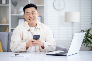 Obraz na płótnie Canvas A happy Korean man is sitting in a bright living room at a table in front of a laptop, holding a phone in his hands, tapping on the screen, texting, smiling and looking at the camera.