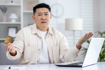 Confused asian man sitting in light office at table in front of laptop, holding card in hand, upset about failed online shopping, broken computer, failed online transaction.