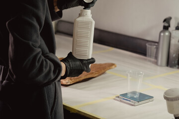 A woman in black protective gloves takes a white bottle with a primer in the workshop. Unscrews the bottle cap. There is a leather bag in the background