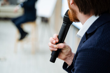 A speaker holds a black microphone in his hand, a person makes a speech on stage, a lecture with a microphone, an object that transmits a voice.