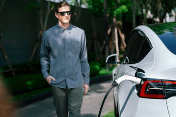 Young man travel with EV electric car charging in green sustainable city outdoor garden in summer...