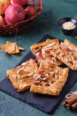 Sliced open apple pie on puff pastry with apple slices, sprinkled with almonds, on a black slate board on a green concrete background. Recipes apple pie