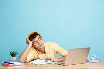 Attractive tired boy student sitting at desk using laptop looking away isolated on blue background