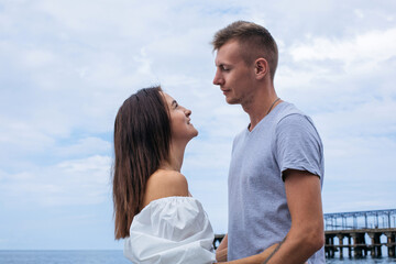 Young couple face to face, holding hands, standing on the shore of the beach in profile by the sea