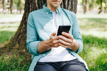 Happy young handsome man sitting on the grass outdoors and using smartphone