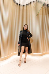 A beautiful model in a short dress and a leather trench coat, posing at a fashion city photo shoot, walking around the city.