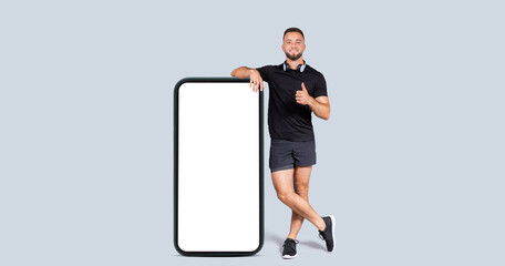 Glad millennial caucasian man athlete in sportswear, headphones show thumb up hand sign near phone with empty screen