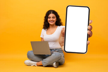Excited indian lady sitting on floor using pc, showing phone