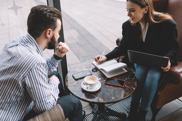 Cheerful young coworkers discussing project in cafe using laptop and taking notes