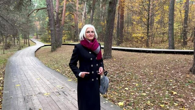 Peaceful senior woman with gray hair walks in forest, admires nature. Beautiful elderly female model in black coat enjoying tranquility. Harmony with nature, and solitude.