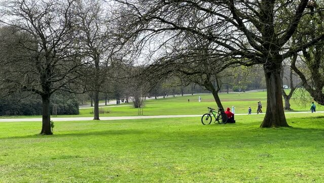 Early spring scene in the city park. Man with bicycle sitting by tree in the Heaton Park, Manchester, UK, family walking past.  Unrecognizable people.