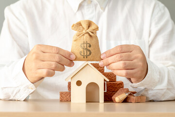 Businessman hand holding the money bag and home model and brick put on the desk in the office, Loan...