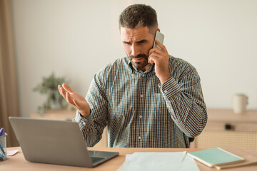 stressed man talking on cellphone managing tasks on laptop indoor