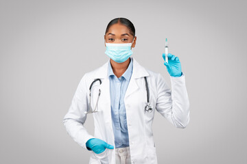 Smiling millennial black lady doctor in white coat, protective gloves, mask show syringe