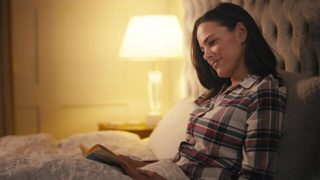 Woman at home lying in bed at night wearing pyjamas reading book - shot in slow motion