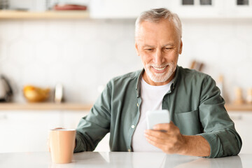 Portrait Of Smiling Senior Man Using Smartphone And Drinking Coffee In Kitchen